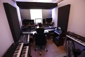 well equipped music studios with sound treatments on the walls and celing, multiple keyboards and a guy sitting at a desk making electronic music on a computer