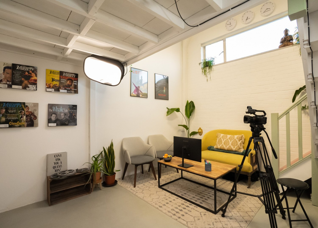 Professional content creation studio for interviews or podcasts, featuring camera, softbox light, seating area, and framed entertainment covers.